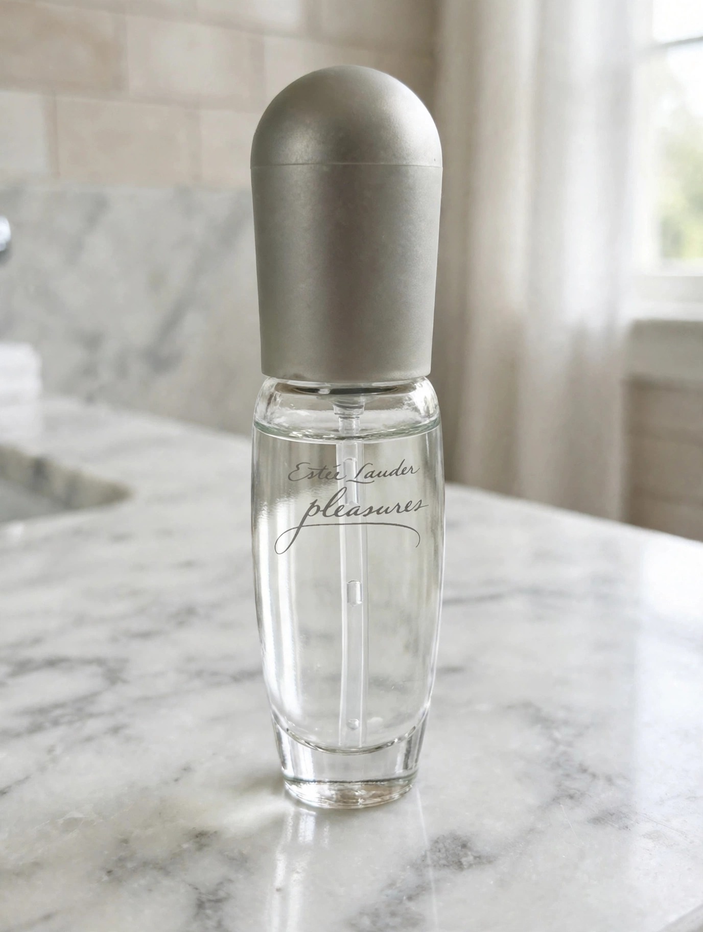 The Estée Lauder Pleasures eau de parfum bottle with its matte silver dome cap standing on a marble bathroom counter